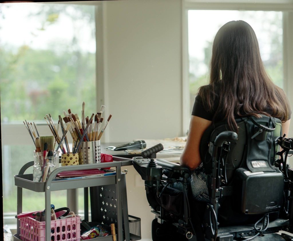 A photo of Reveca from behind. Her arm is resting gently on her armrest, and her straight brown hair is slightly draped over the top of her wheelchair backrest. She is sitting at an art desk in a white room with big windows facing a tree-lined yard. Next to her there is a metal cart with three cylindrical containers full of paint brushes fanning out of the tops and a pink basket of art supplies on the second shelf of the cart. Photograph by Genesis Falls.