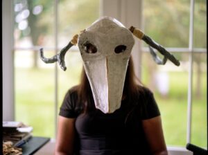 A brown-skinned woman in a black T-shirt photographed from the torso up. Her arms are down at her sides and she has straight, warm brown hair down to her chest. A large mask is covering her face. It is made of grayish-white papier-mâché with two dark antlers pointing down and out to the sides. There is a corner of a desk to the left of her, showing a glimpse of some piles of notebooks and textiles. The background is blurry, showing large windows with bright green grass and tree tops, outside the room where the figure is sitting. Photograph by Genesis Falls.