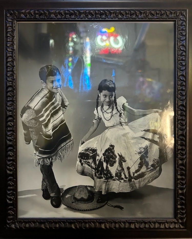 2026 Calaveras neon sign reflected in the framed photo of Ricardo Sepeda’s 8-year-old-mom and uncle posing for a photo at the Club Guadalupano event in Elgin, 1961. Photo credit: Basia Krupa, courtesy of Ricardo Sepeda.