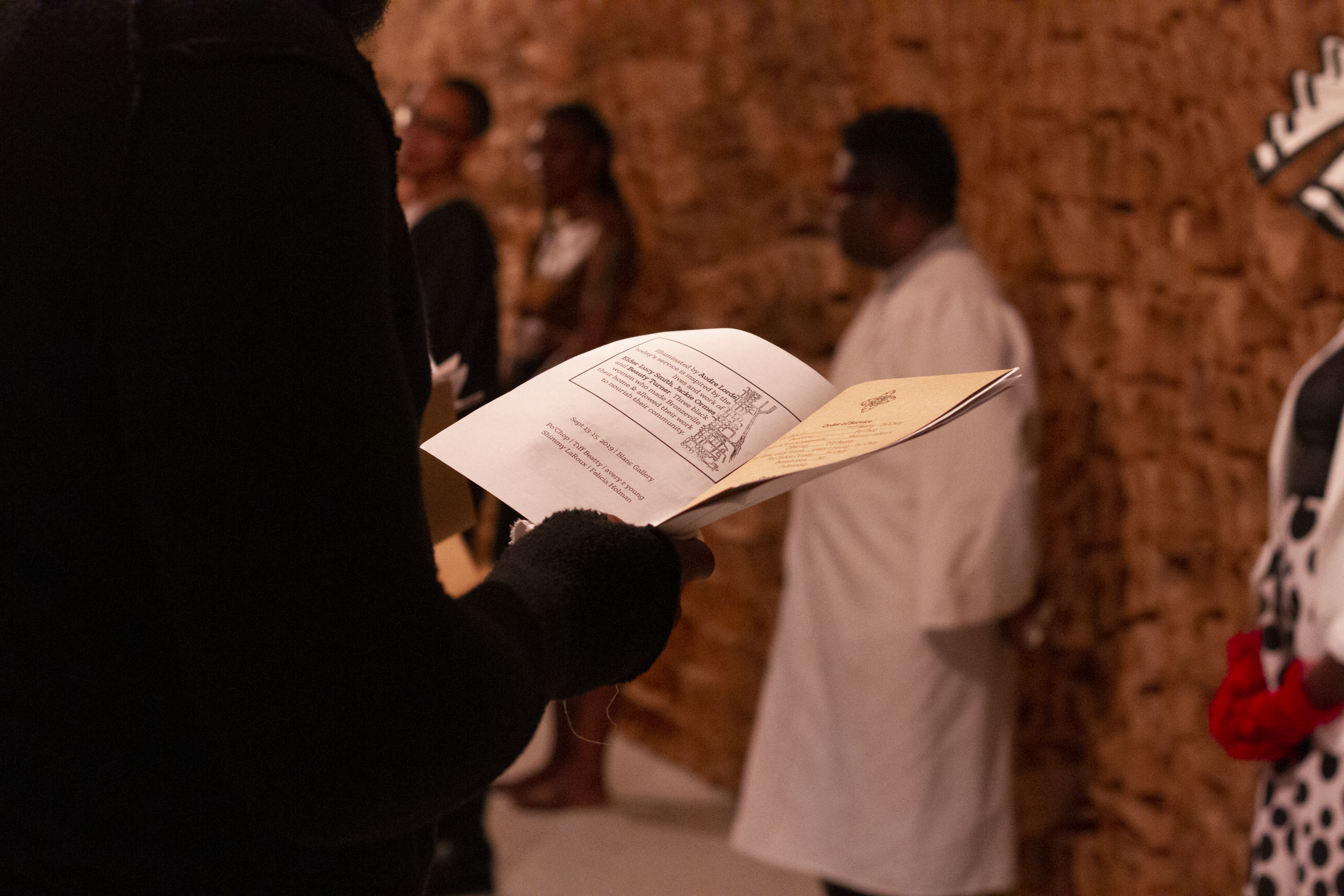 A photo of an audience member holding a program from The People’s Church of the G.H.E.T.T.O with Tiff, Jenn, and avery standing in the background. Photo by Candice Majors.