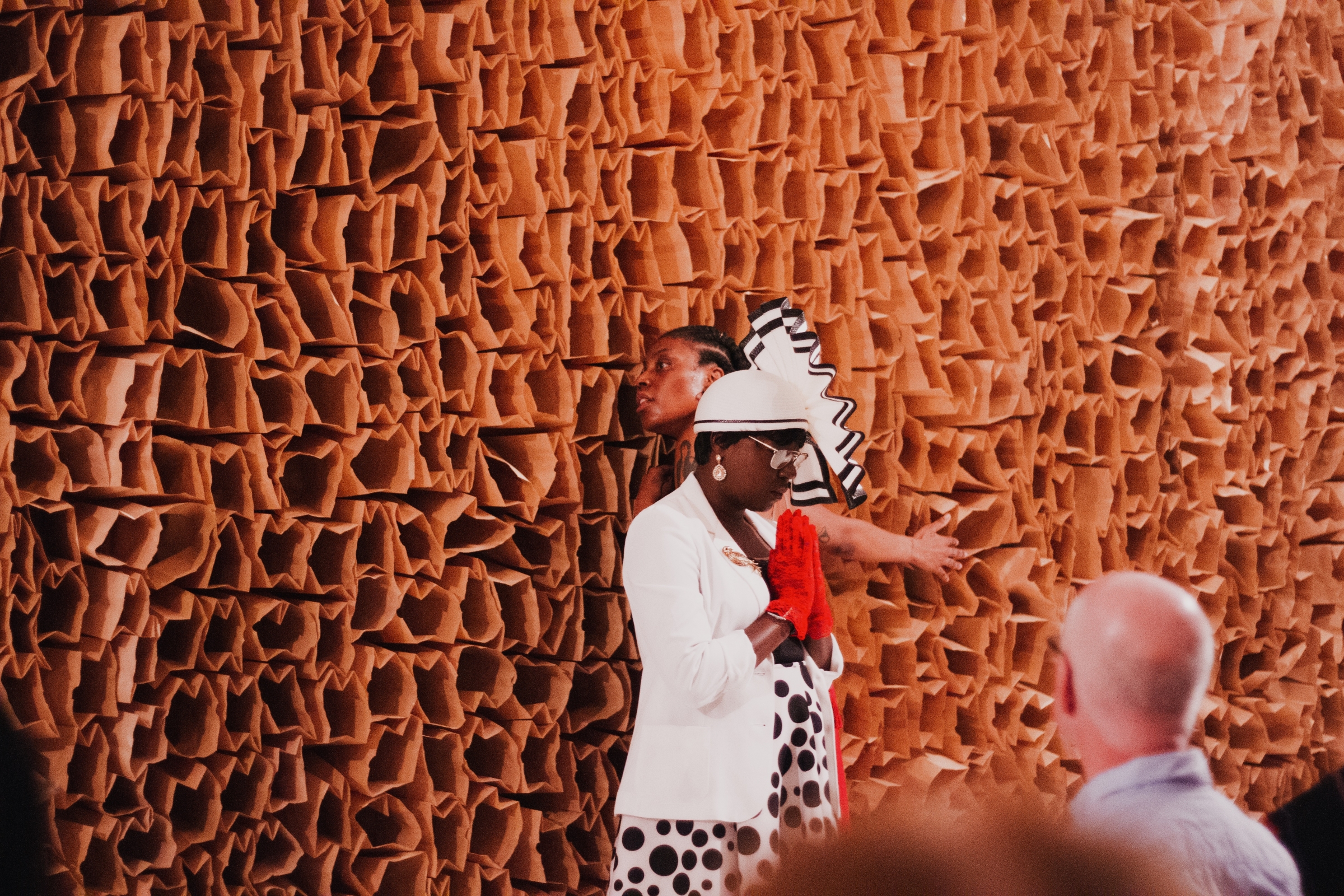 A photo of Shimmy LaRoux, a member of the usher board for The People's Church, standing in front of a brown wall covered in paper bags and an audience, wearing all white with red gloves and palms together in front of her in prayer. Photo by Candice Majors.