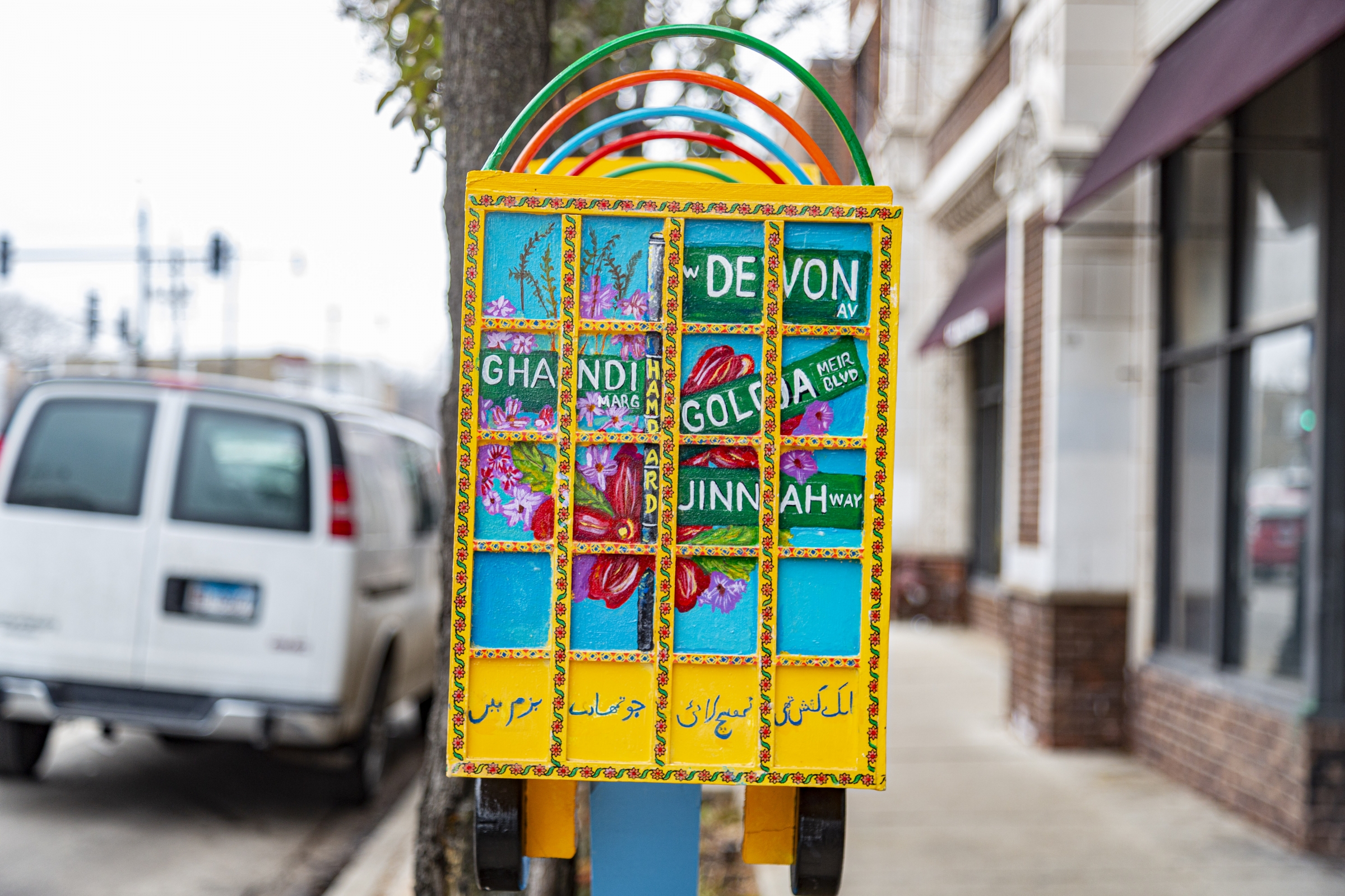 Urooj Shakeel's Truck Art Meets Little Free Library, installation view along Devon Avenue in Chicago. The image shows a small, yellow truck sculpture with bright multicolored images painted on each side, held up by a bright blue stake in the ground. Photo by Mark Blanchard.